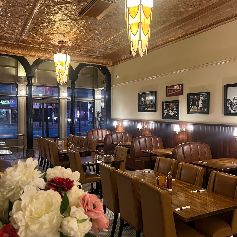 Elegant restaurant dining room with leather chairs, wooden tables, ornate tin ceiling, vintage photographs, and glowing Art Deco pendant lights.