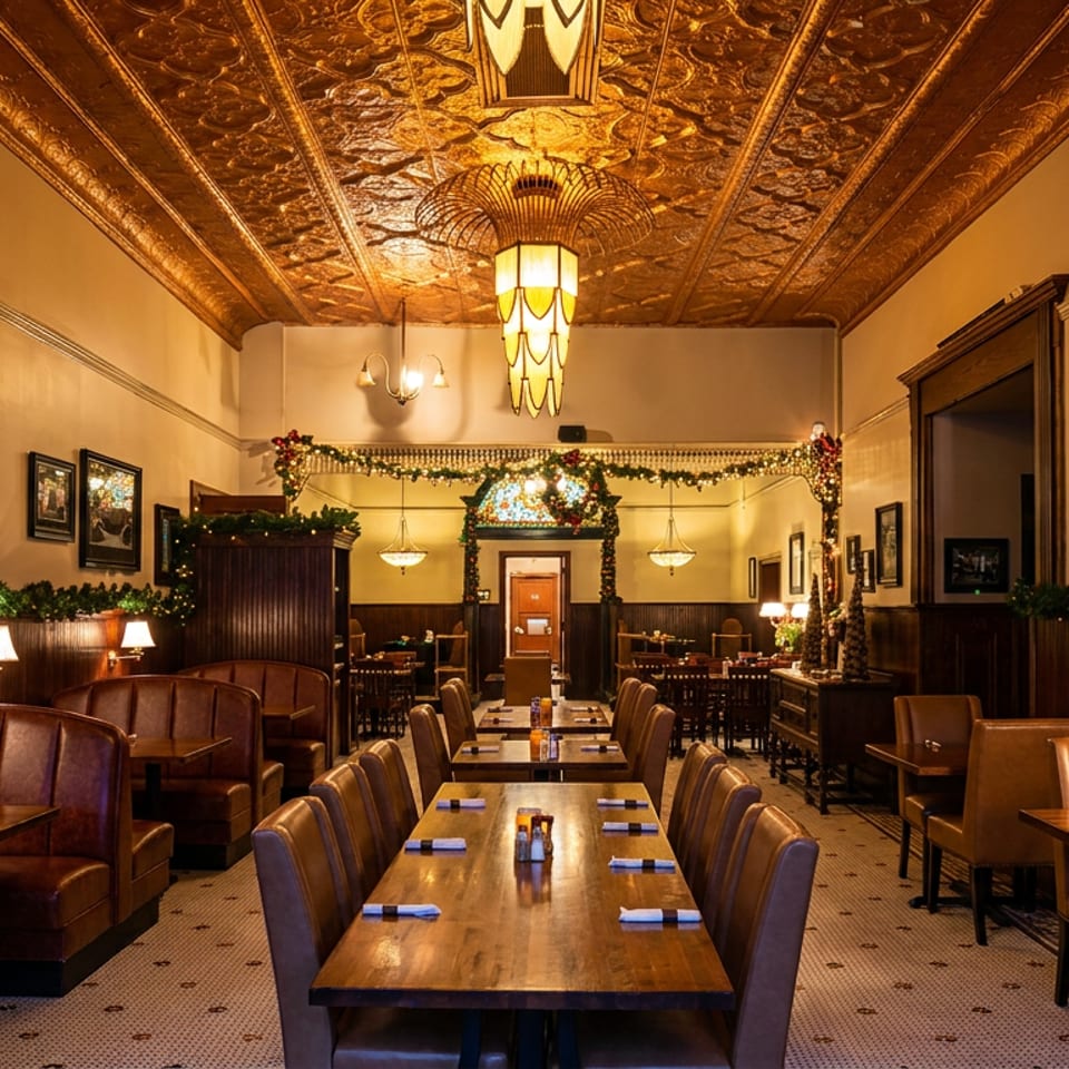 Warmly lit restaurant dining room with leather booths, wooden tables, vintage framed photos, ornate gold ceiling, and festive garlands.