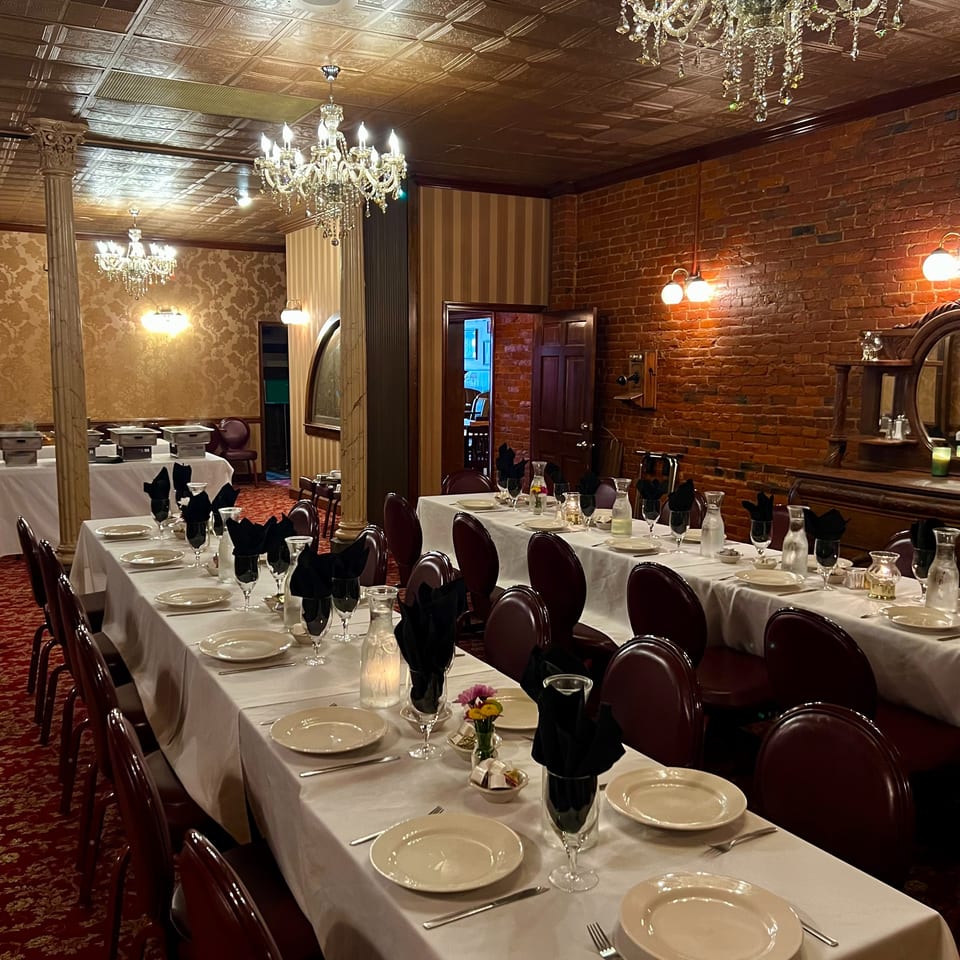 Elegant restaurant dining room with white tablecloths, black napkins, crystal chandeliers, exposed brick walls, and ornate tin ceiling.
