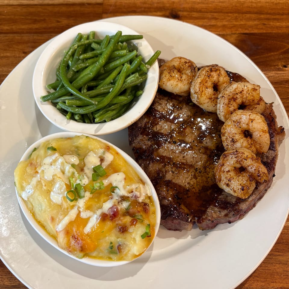 Grilled steak topped with shrimp, served with green beans and loaded cheesy potatoes on a white plate.