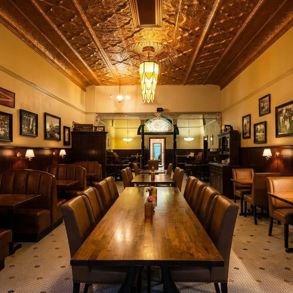 Warmly lit historic restaurant dining room with leather booths, wooden tables, framed photographs, and an ornate pressed-tin ceiling.