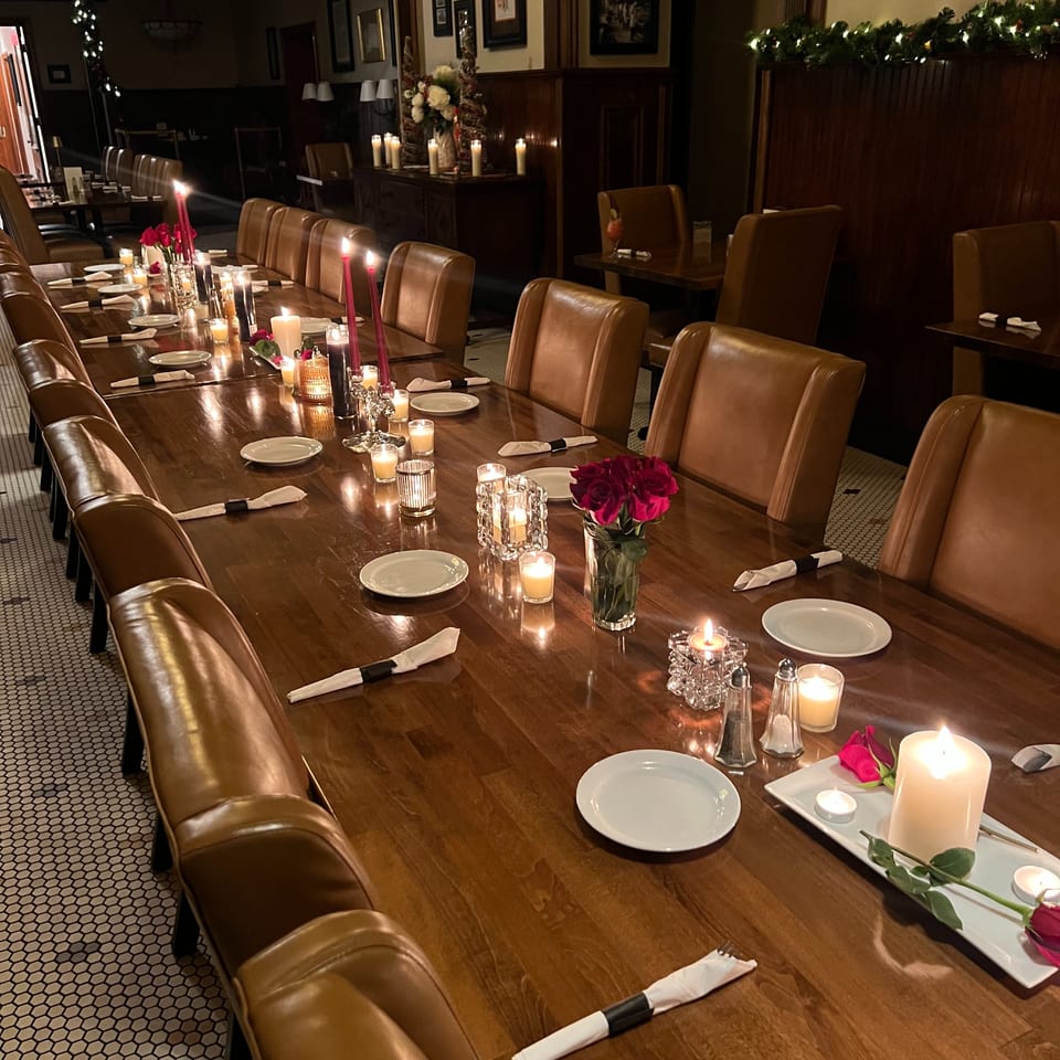 Elegant restaurant dining room with candlelit wooden tables, leather chairs, white place settings, red roses, and festive garlands.